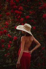 Outdoor fashion portrait of elegant beautiful woman wearing luxury wide brim straw hat, red dress with naked back, posing near blooming roses
