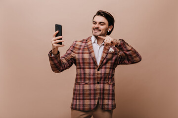 Elegant young brunette in white shirt, plaid brown jacket, with earphones, looking at phone and...