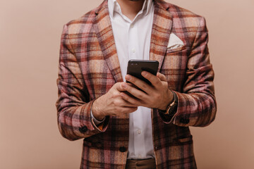 Fototapeta premium Faceless photo of elegant man in white shirt and plaid brown blazer, holding phone and posing isolated on beige background