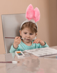 A little girl in cute headband with pink bunny ears diligently draws with a brush with watercolor...