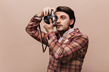 Anxious young dark-haired photographer in plaid blazer holding camera and looking aside. Man posing isolated over beige background