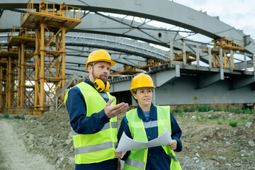 Construction workers in reflective clothing and work helmets looking at building and examining blueprint on construction site
