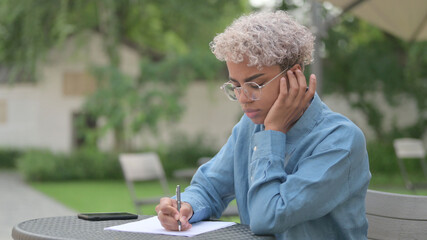 African Woman Thinking while Writing on Paper in Outdoor Cafe