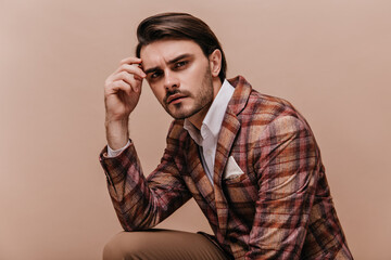 Portrait of thinking young man with brunette hair and bristle, wearing white shirt, plaid blazer, looking into camera and holding hand near head, isolated on beige background