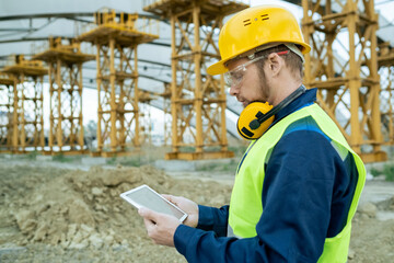 Architect in work helmet examining blueprint on digital tablet while working on construction site outdoors