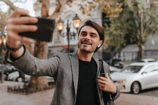 Portrait of lovely young gentleman in black t-shirt and plaid vintage blazer, making selfie against city landmark background and smiling