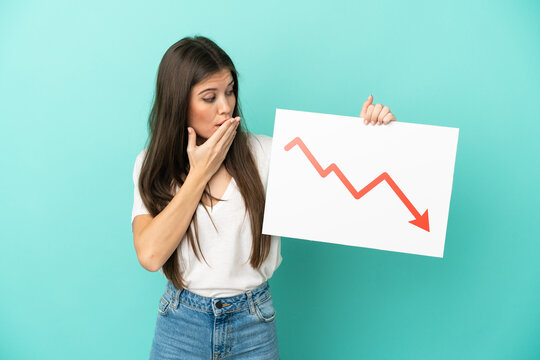 Young caucasian woman isolated on blue background holding a sign with a decreasing statistics arrow symbol with surprised expression
