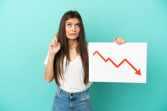 Young caucasian woman isolated on blue background holding a sign with a decreasing statistics arrow symbol with fingers crossing