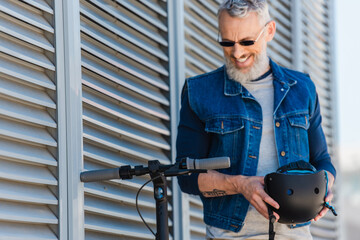 bearded middle aged man in sunglasses smiling and holding helmet near electric scooter