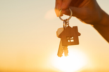 The hand holds the key to the new house with a keychain in the form of a house on the background of the sunset. - obrazy, fototapety, plakaty