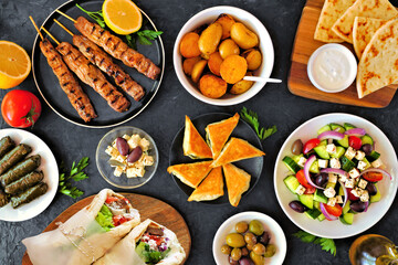 Greek food table scene, top down view on a dark background. Souvlaki, gyros wraps, salad,...