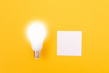 A Power Saving Lamp with White Note Paper Lying on Yellow Table, Blank Template with Copy Space - Top View, Flat Lay