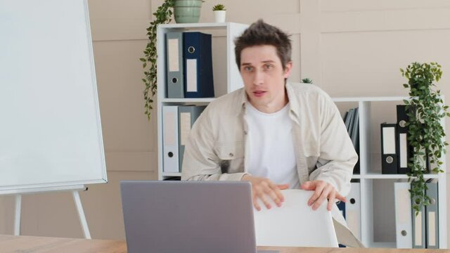 Funny caucasian man male millennial freelancer sitting down at desk in home office opens laptop screaming with fear scared frightened by image on screen hiding behind chair in horror closes computer