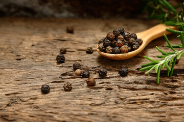 Black pepper in a wooden spoon with branch rosemary on a weathered wooden floor as background.