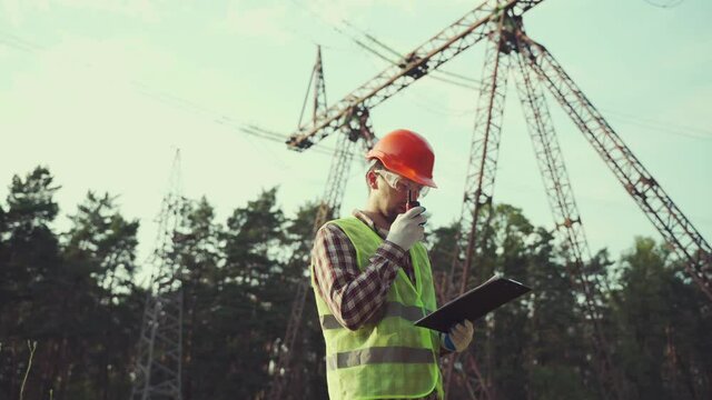 Caucasian engineer in protective uniform and orange hard hat holds walkie-talkie and clipboard on background of electric transmission line. Electrical worker with radio set near high voltage tower.