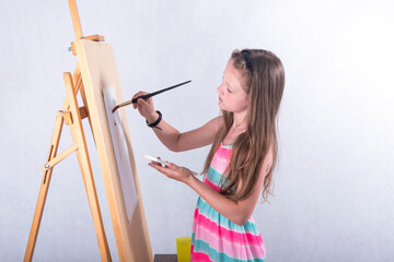 A beautiful girl-artist of school age in the studio on a white background with a brush in her hand and a watercolor inspiringly draws a picture behind an easel on paper