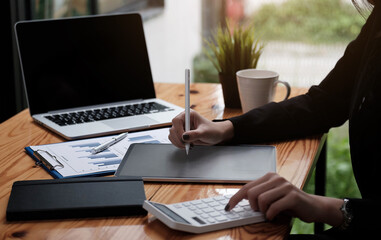 Fototapeta premium Business woman or accountant hand using calculator, working on taplet and laptop computer with documentary data graph paper on the table at modern home office. Online marketing concept.