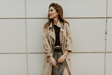 Portrait of attractive lady in checkered midi skirt and beige trench coat looks away and poses with retro camera outside.