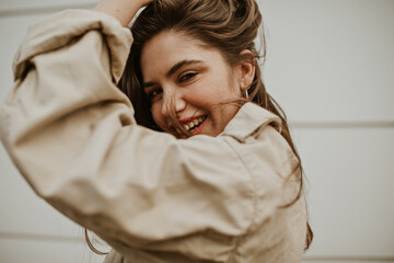 Brown-eyed woman in beige trench coat ruffles hair outside. Happy girl in jacket smiles and poses in good mood.