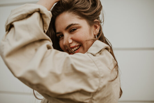 Brown-eyed Woman In Beige Trench Coat Ruffles Hair Outside. Happy Girl In Jacket Smiles And Poses In Good Mood.