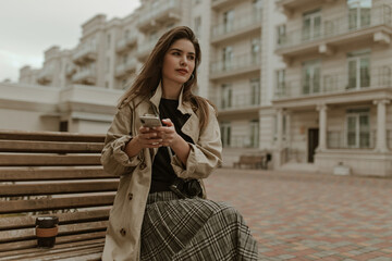 Attractive woman in black top, checkered skirt and beige trench coat sits on bench outside and holds phone. Girl in stylish outfit looks away.