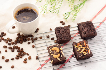 4 square dark brownies with chocolate chips, almonds and nuts on a baking rack, 1 white cup of coffee and coffee beans placed on a white cloth.
