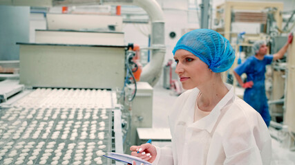 Candy factory. Controller checking conveyor with candies. Young woman in uniform holding folder and inspecting conveyor belt with fresh candies in confectionery factory.