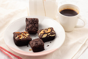 4 square dark brownies with chocolate chips, almonds and nuts on a white plate, coffee beans placed on a white cloth. sweets for tea time, coffee time, or break time.