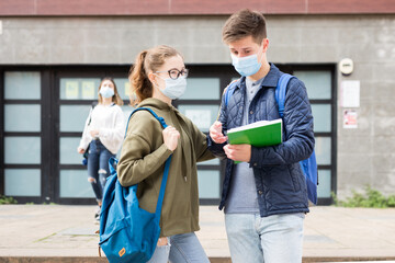 Two teenage students in protective masks discuss homework near the college building on a sunny day