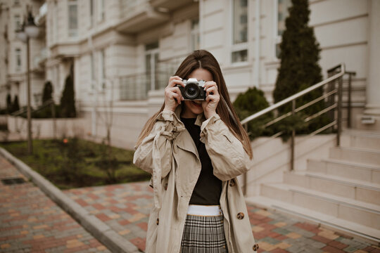 Brunette young woman in plaid skirt and beige trench coat walks outside and take photos at retro camera.