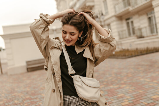 Joyful brunette woman in black top, checkered skirt and trench coat touches hair, smiles and walks outside in good mood.