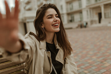 Brunette charming woman in beige trench coat and black t-shirt smiles sincerely, sits on wooden bench and takes selfie outside.