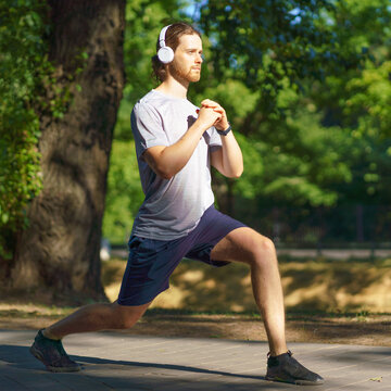 Full length of young male sportsman in headphones working out in green park in sunny summer day, active sportive man dressed in activewear doing sports exercises lunges outdoors