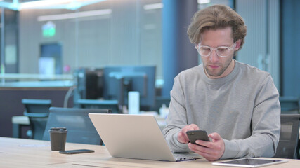 Young Man with Laptop using Smartphone at Work