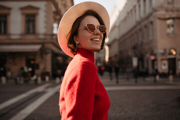 Optimistic beautiful lady in beige hat and eyeglasses smiles outside. Charming woman in red sweater...