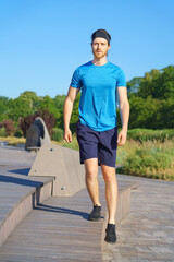 Full length shot of young athletic guy in sports clothes doing exercises on wooden stairs outdoors, sportive active man training in sunny morning. Urban sport and workout concept