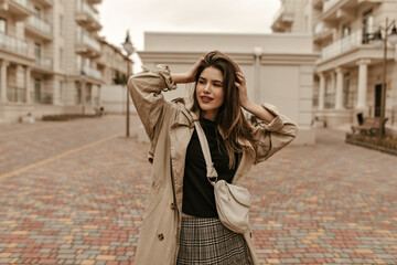 Beautiful brunette woman ruffles hair and smiles gently. Pretty girl in trench coat and checkered skirt walks outside.