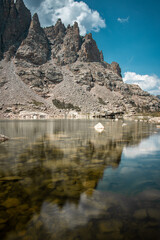 View of sky pond in Rocky Mountain National Park in Colorado. Sharp teeth-like peaks can be seen as well as the pond water with a reflection of the mountains above and blue skies with clouds. 