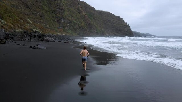 Drone clip flying over a young man jogging on a tropical beach with black sand in Playa de Nogales, La Palma, Canary Islands in Spain
