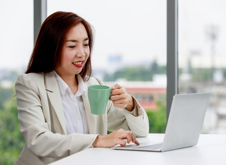 Content young Asian female entrepreneur in stylish suit drinking coffee and using laptop while working on business project in modern workspace