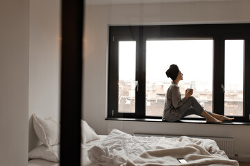 Woman in grey pajamas and towel on head sits on windowsill and enjoys city view. Beautiful lady holds tea cup in bedroom.