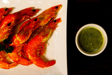 Top view shot of Fresh delicious tasty cooked grilled roasted seasoning orange sea tiger shrimps decorated with parsley on white dish with green spicy seafood sauce on old dark brown wooden table