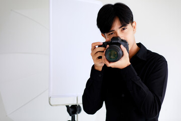 Asian handsome male photographer wearing casual black shirt, holding a camera, posing and sitting in a studio with light equipments.