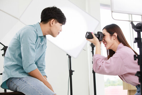 Asian friendly confident long brown hair female photographer smile hold dslr camera in hands try to closeup shoot handsome male model who sit on chair lean forward close to lens in photography studio