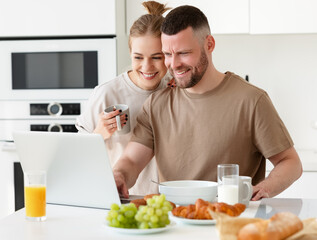 Fototapeta na wymiar Young beautiful happy family couple reading news on laptop during morning breakfast at home