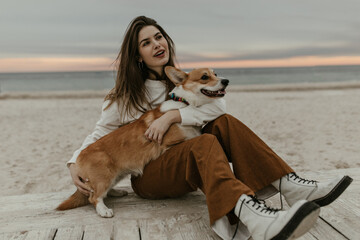 Charming brunette woman in beige sweatshirt and brown pants sits at beach, looks away and plays with corgi.