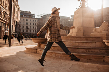 Pretty cool woman jumps near statue in old European city square. Charming girl in checkered coat...