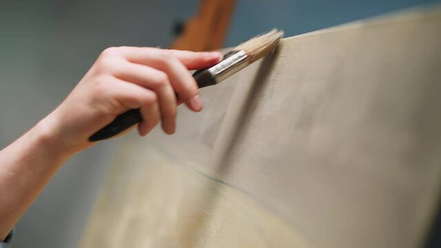 Slow motion, creative person at work, a talented adult man artist covers the canvas with beige oil paint, priming and draws the background, close-up view of the hands.