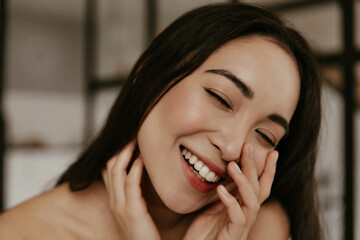 Good-humored brunette Asian woman smiles with closed eyes and touches her face skin. Portrait of pretty joyful lady in apartment.