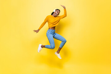 Cheerful energetic young African American woman wearing headphones jumping and listening to music on yellow isolated studio background